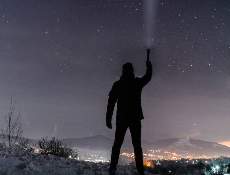 Man in outdoors at night pointing flashlight to the sky.  This flashlight symbolizes the usefulness of a hand crank solar powered flashlight for survival gear enthusiasts at night or during an emergency.